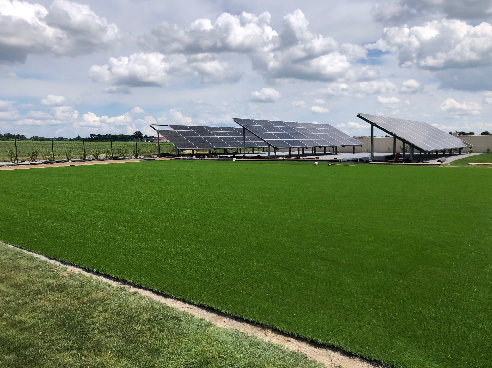 Instalacja paneli słonecznych na zielonym trawniku, widoczne panele ustawione w rzędach, częściowo zacienione, błękitne niebo z białymi chmurami w tle.