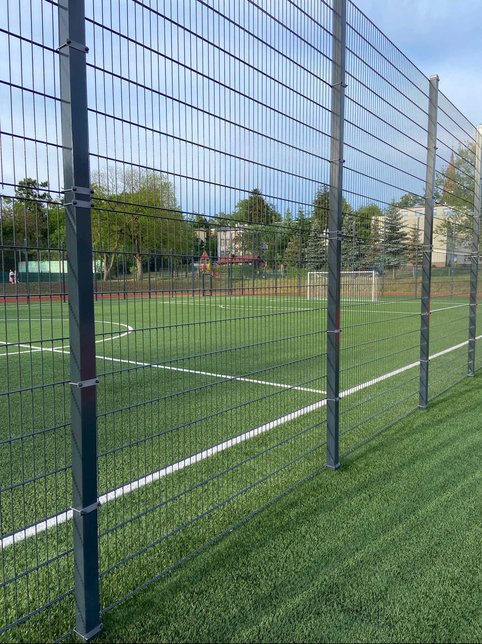 Ogrodzenie panelowe boiska sportowego z zieloną sztuczną nawierzchnią i widoczną bramką w tle, słupki mocowane na metalowe obejmy.