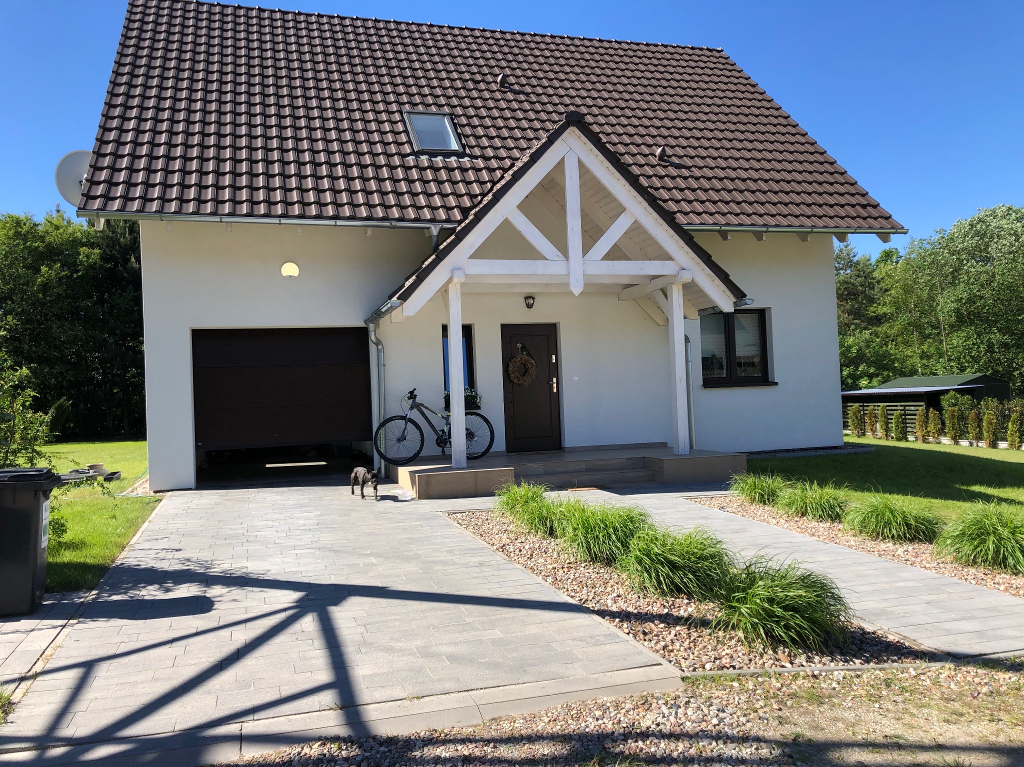 Biały dom jednorodzinny z brązowym dachem, podjazd z szarej kostki brukowej, rower oparty o wejście, mały pies na podjeździe.