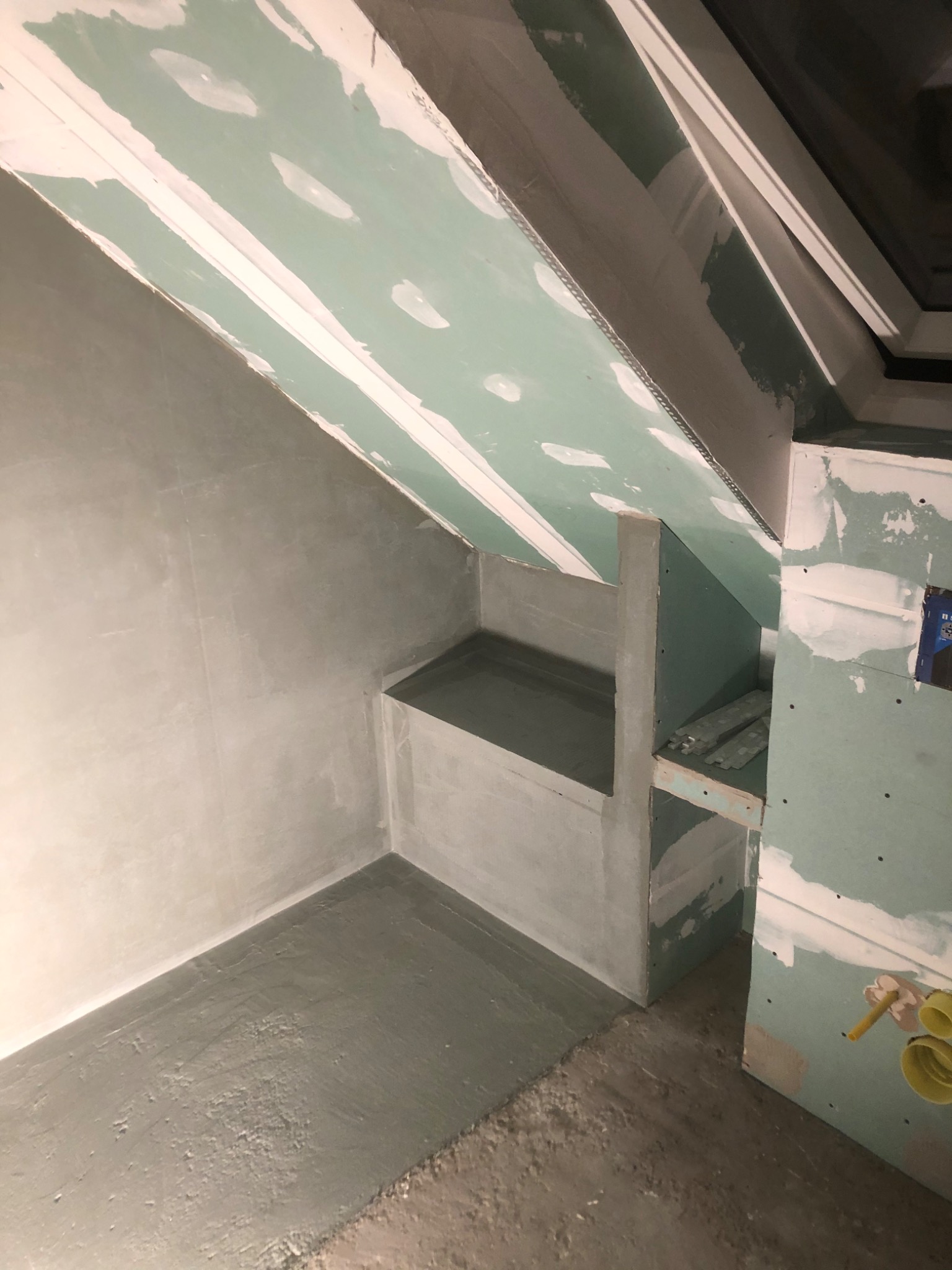 Łazienka na poddaszu w trakcie prac wykończeniowych: ściany pokryte szarą warstwą, sufit z płyt gipsowo-kartonowych, wnęka z półkami w stanie surowym, widoczne elementy instalacji hydraulicznej.