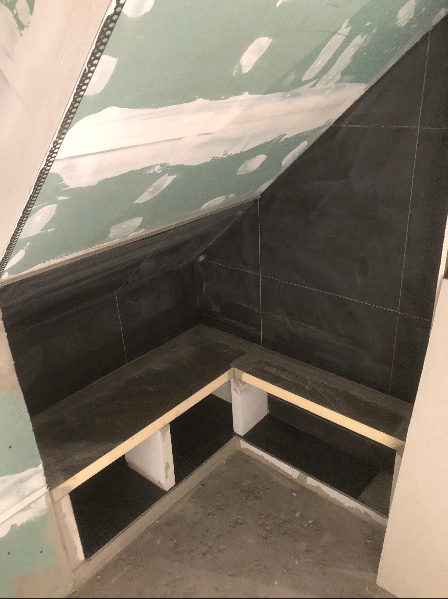 Niedokończona łazienka na poddaszu: czarne płytki na ścianach, surowy gips na skosach, konstrukcja półek z płytek i drewnianych belek, surowa podłoga.