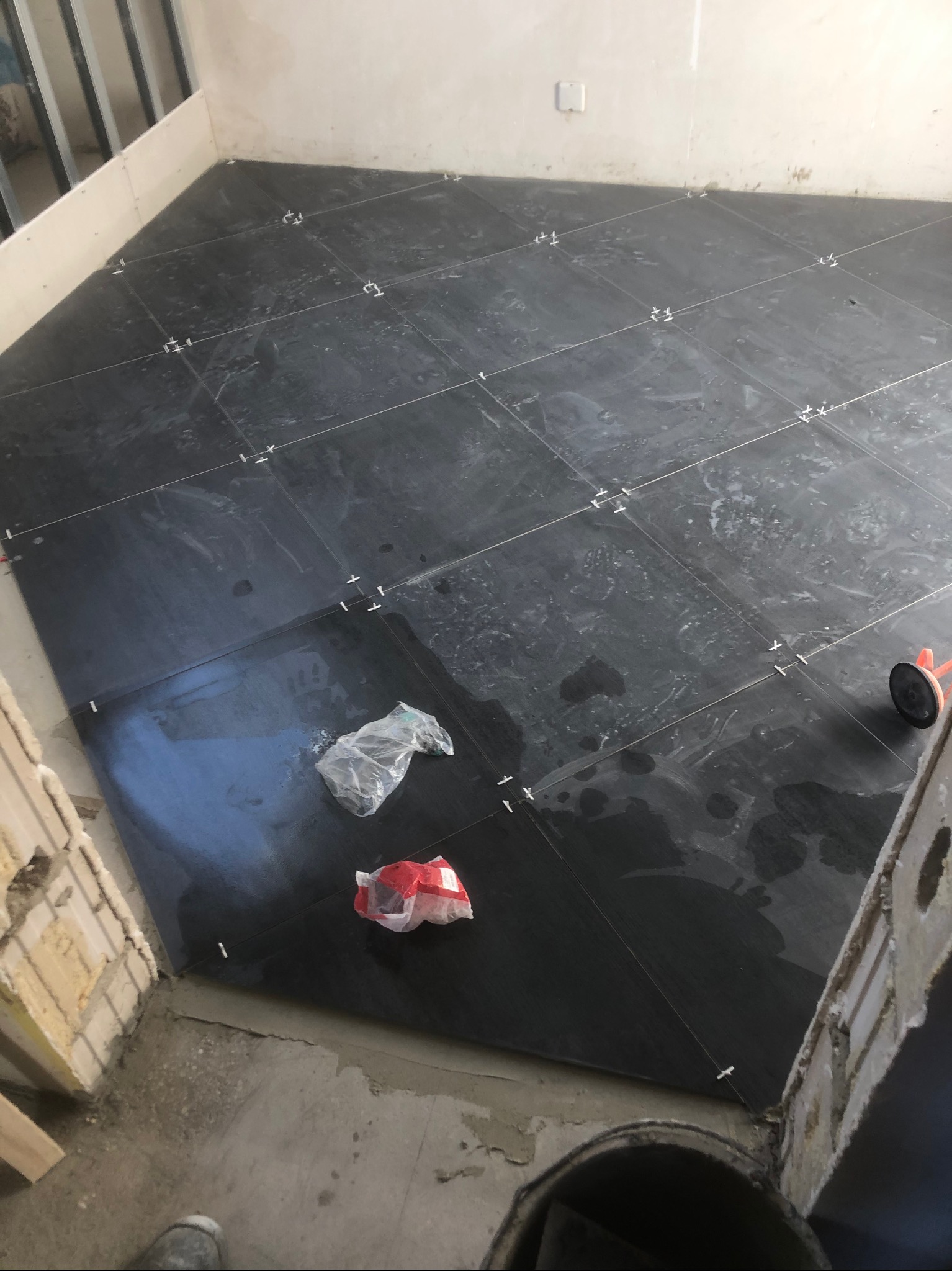 Układanie ciemnych płytek podłogowych z krzyżykami dystansowymi, widoczne resztki zaprawy i narzędzia glazurnicze, etap prac wykończeniowych.