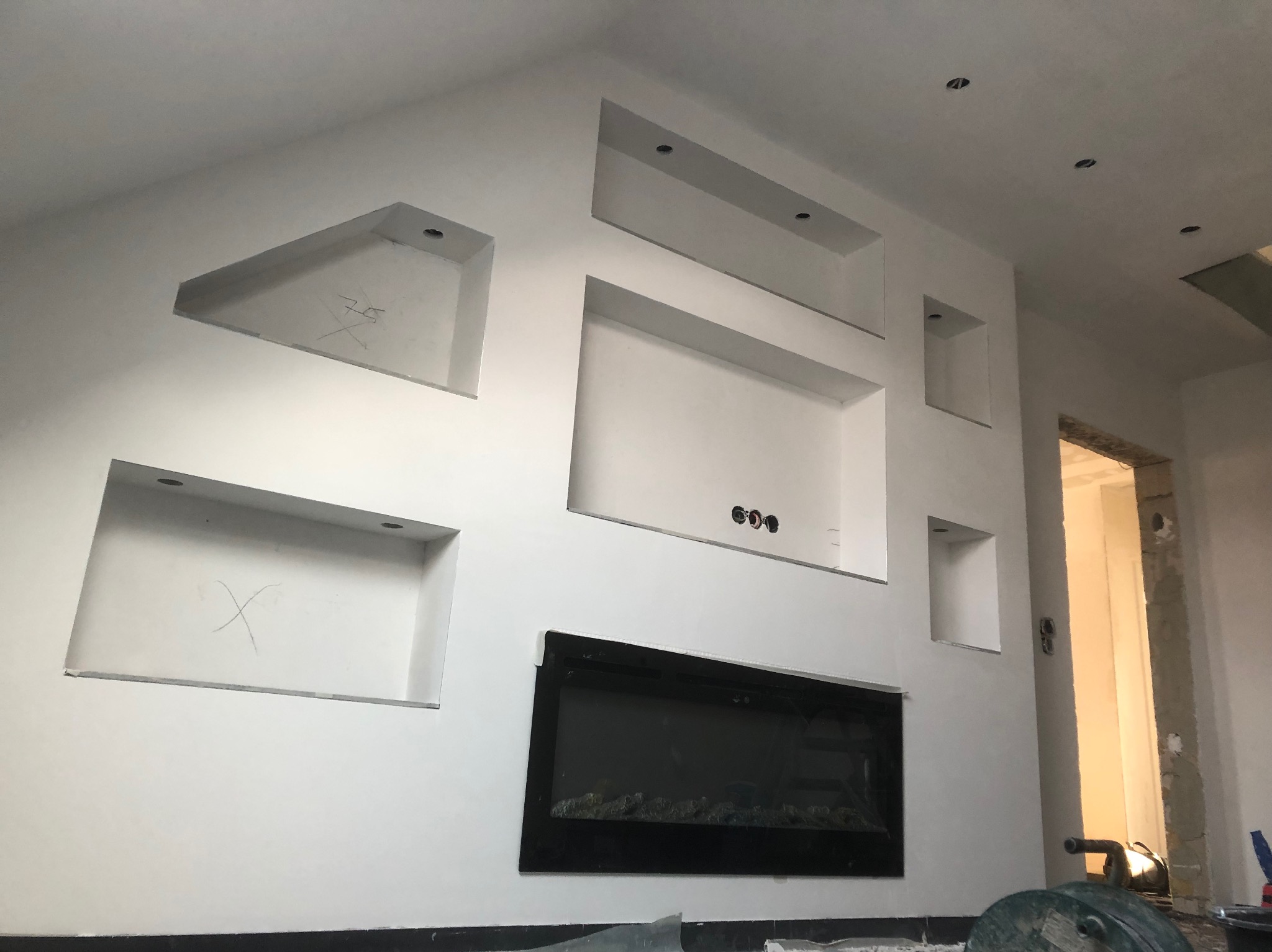 Ściana po gładzi z wnękami o różnych kształtach i rozmiarach, przygotowana pod montaż oświetlenia punktowego i kominka elektrycznego, widoczne ślady ołówka na ścianie.