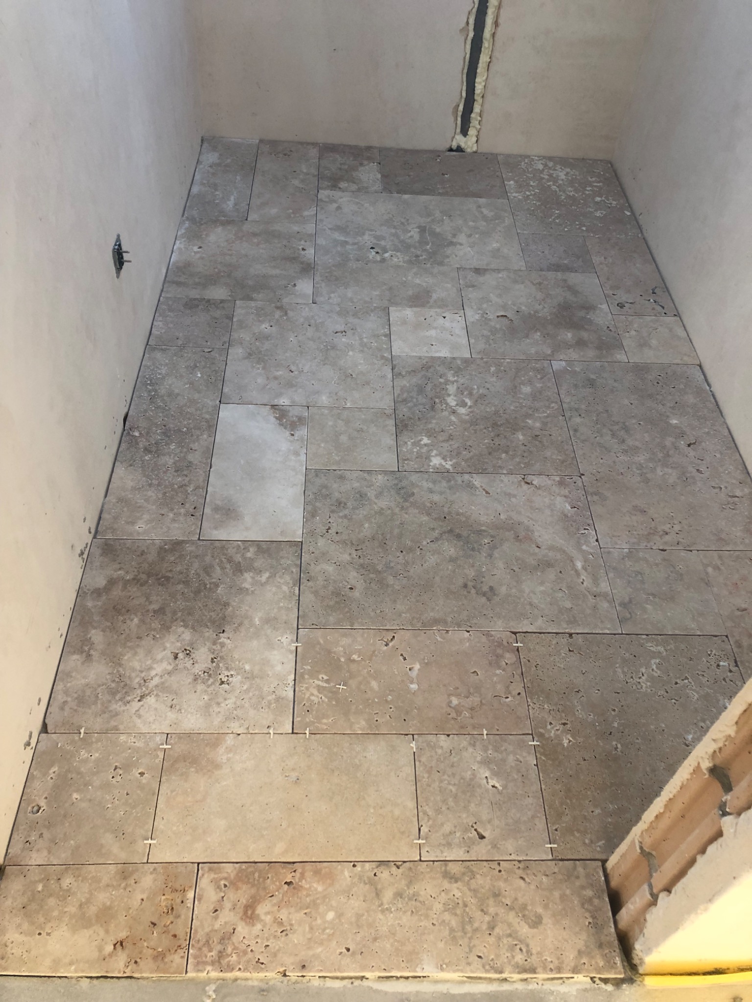 Układanie beżowych płytek trawertynowych na podłodze w trakcie remontu łazienki, widoczne krzyżyki dystansowe i fragment surowej ściany.