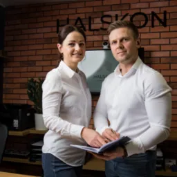 Dwoje uśmiechniętych pracowników w biurze, przeglądających dokumenty na tle ściany z cegły i logo firmy.