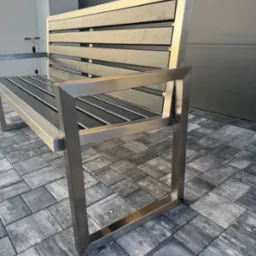 Nowoczesna ławka parkowa z nierdzewnej stali z poziomymi listwami siedziska i oparcia, na szarej kostce brukowej, z widoczną ścianą budynku w tle.