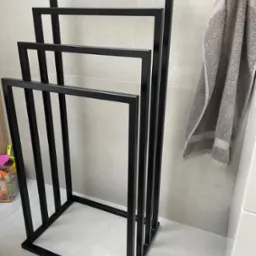 Czarny, metalowy stojak na ręczniki w łazience z szarym ręcznikiem wiszącym obok, widoczne detale wykończenia i minimalistyczny design.