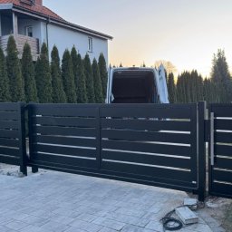 Mat-Spaw Mateusz Nóżka - Nowoczesna, przesuwna brama wjazdowa z poziomymi szczeblami, pomalowana na ciemny kolor, z widocznym przewodem elektrycznym i fragmentami kostki brukowej w trakcie układania, na tle domu...