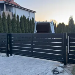 Nowoczesna, przesuwna brama wjazdowa z poziomymi szczeblami, pomalowana na ciemny kolor, z widocznym przewodem elektrycznym i fragmentami kostki brukowej w trakcie układania, na tle domu...