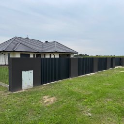Mat-Spaw Mateusz Nóżka - Nowoczesne ogrodzenie panelowe w kolorze antracytowym, z elementami murowanymi, otaczające dom jednorodzinny z ciemną dachówką. Widok z zewnątrz, w tle zielony trawnik i błękitne niebo.