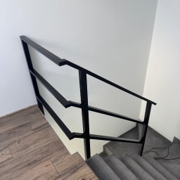 Mat-Spaw Mateusz Nóżka - Minimalistyczna, czarna balustrada schodowa z metalu, widziana z góry. Kontrastujące połączenie drewnopodobnych paneli podłogowych i szarych płytek na schodach.