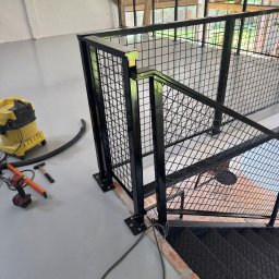 Mat-Spaw Mateusz Nóżka - Czarna, metalowa balustrada z siatką na schodach w loftowym wnętrzu. Widoczne narzędzia i odkurzacz, sugerujące etap montażu. Surowy styl, cegła i drewno w tle.