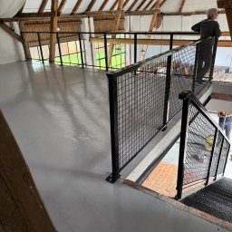 Mat-Spaw Mateusz Nóżka - Antresola z metalową balustradą z siatki w loftowym wnętrzu. Widoczna osoba opierająca się o balustradę i schody. Drewniana konstrukcja dachu.