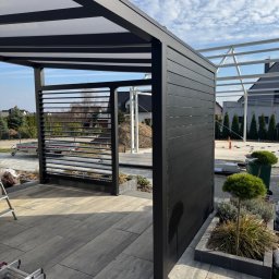 Mat-Spaw Mateusz Nóżka - Nowoczesna, czarna pergola z elementami lamelowymi i przezroczystym zadaszeniem, otoczona kostką brukową i zielenią. W tle widoczny budynek w trakcie budowy.