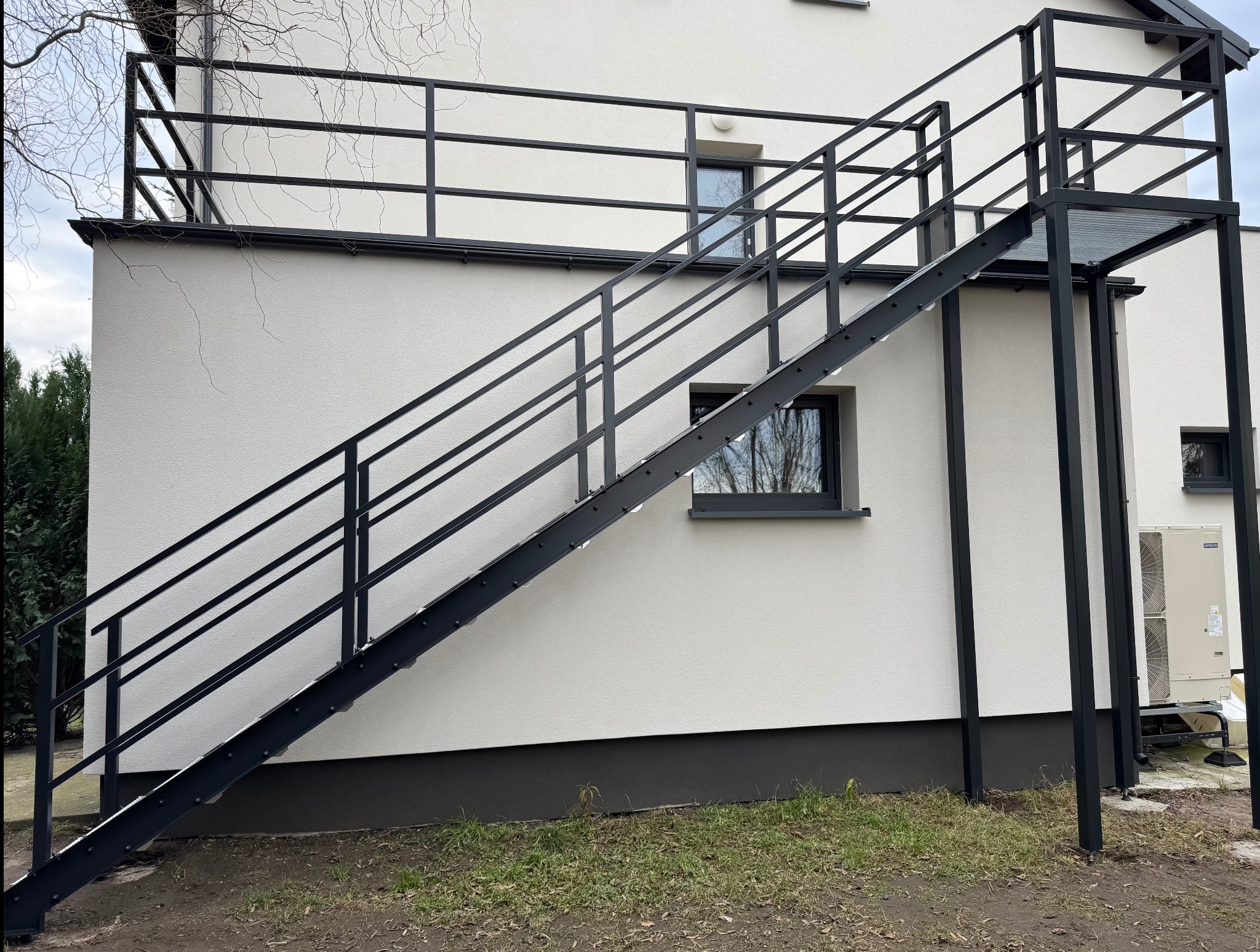 Zewnętrzne schody metalowe w kolorze czarnym, prowadzące na balkon z balustradą, przylegające do białego budynku z widocznymi oknami i klimatyzatorem.