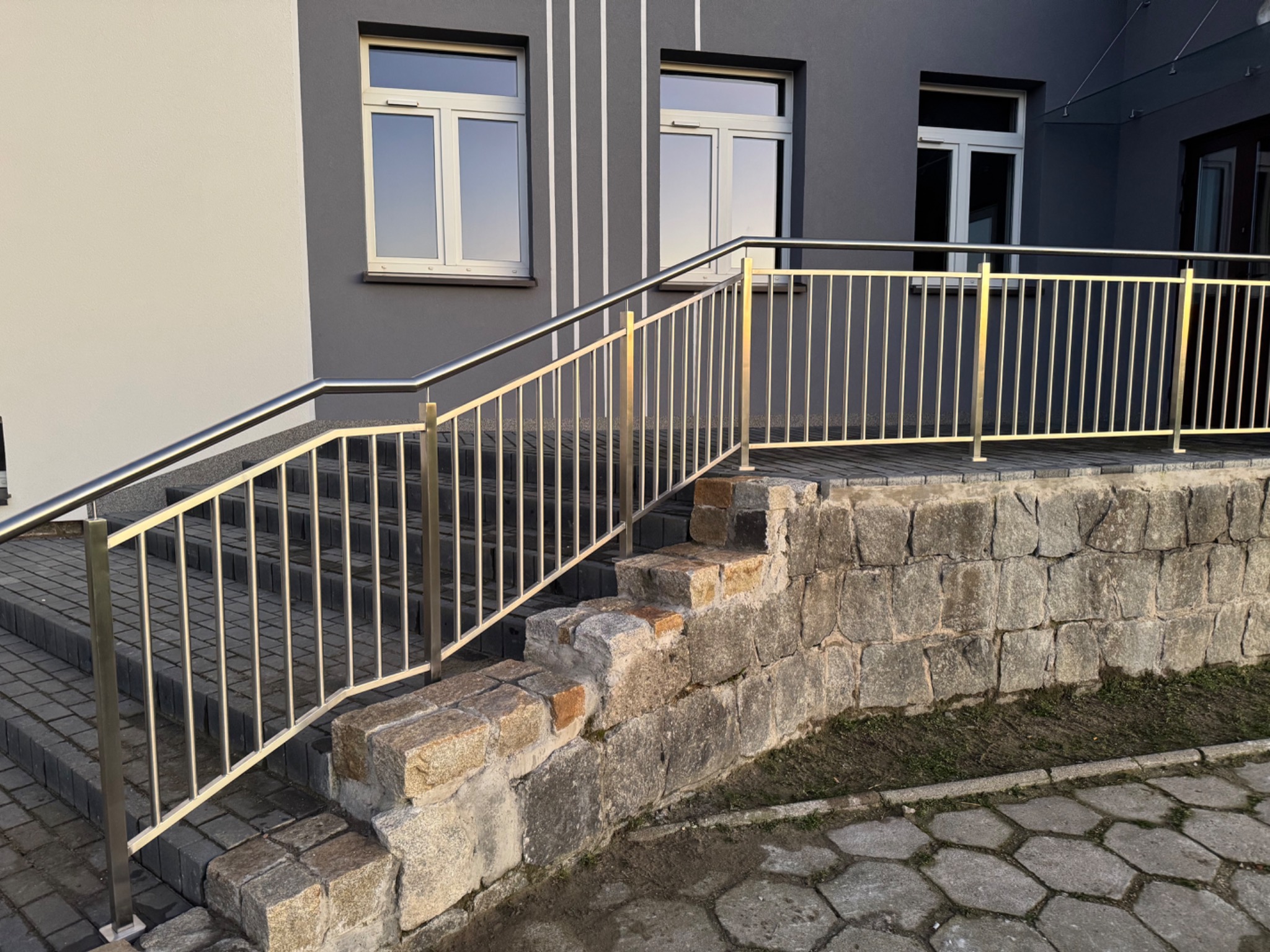 Zewnętrzne schody z szarej kostki brukowej, z balustradą ze stali nierdzewnej o złotym odcieniu, prowadzące do budynku z szarą elewacją i białymi oknami.