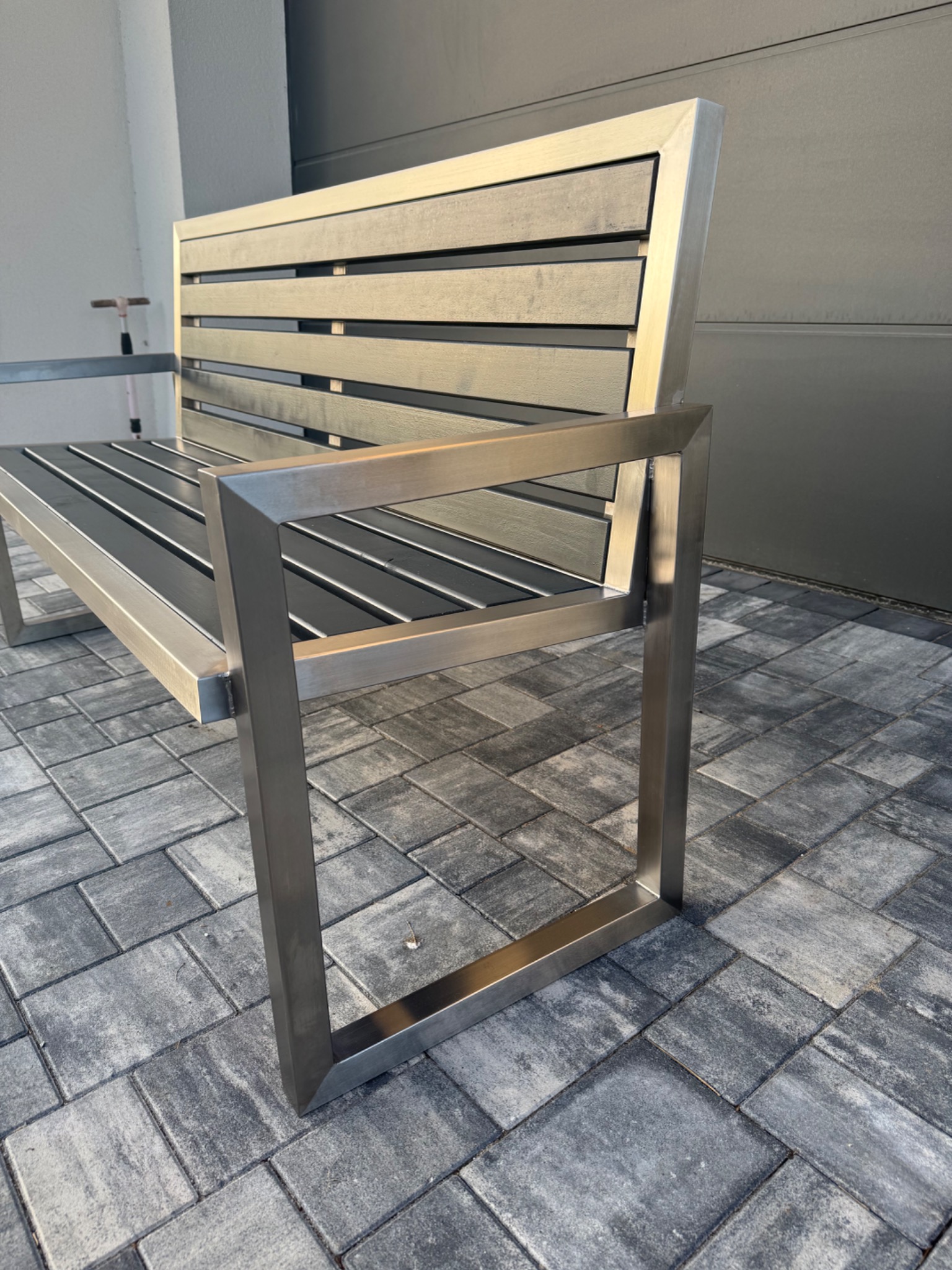 Nowoczesna ławka parkowa z nierdzewnej stali z poziomymi listwami siedziska i oparcia, na szarej kostce brukowej, z widoczną ścianą budynku w tle.