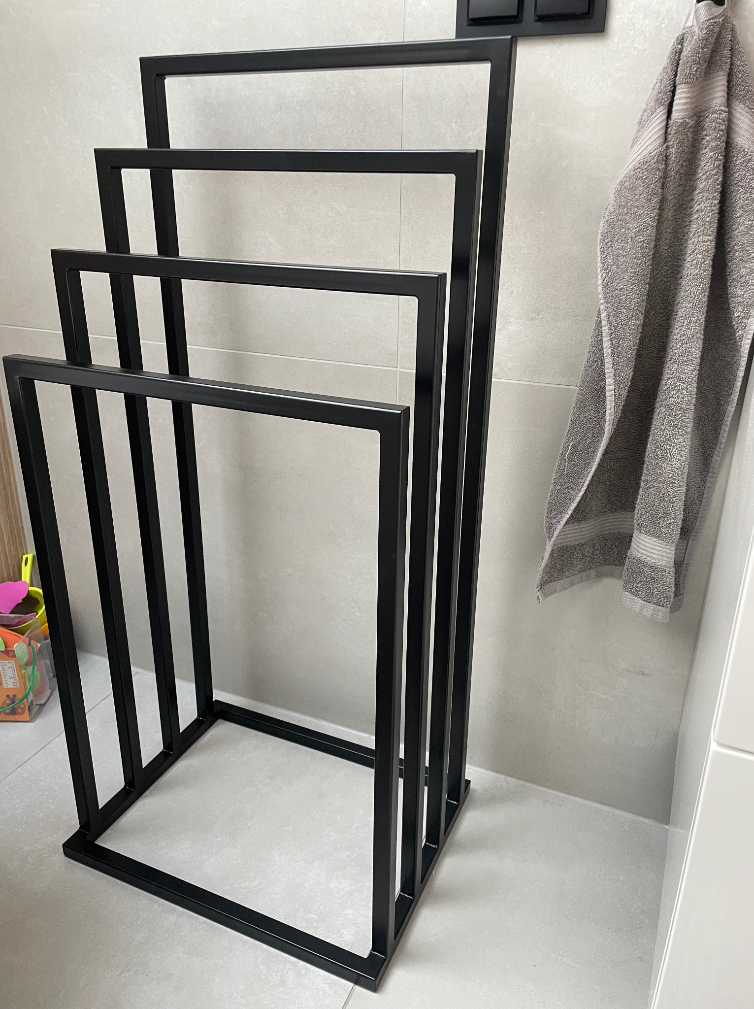 Czarny, metalowy stojak na ręczniki w łazience z szarym ręcznikiem wiszącym obok, widoczne detale wykończenia i minimalistyczny design.