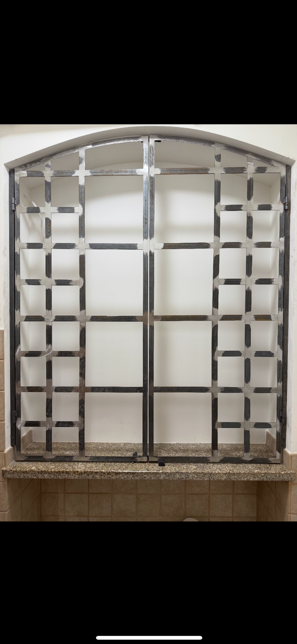 Metalowa witryna okienna z kratą, surowy wygląd spawów, osadzona na blacie z płytek. Widoczne zawiasy i zamknięcie. Minimalistyczny design.