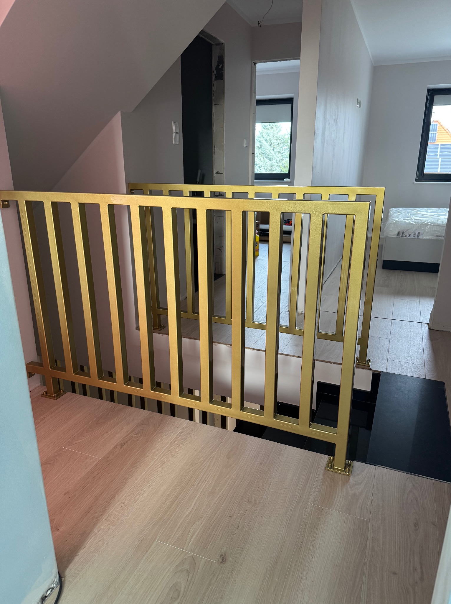 Złota, metalowa balustrada schodowa z pionowymi szczeblami, zamontowana w jasnym wnętrzu domu. Widok z dołu na schody i fragment pokoju z oknem.