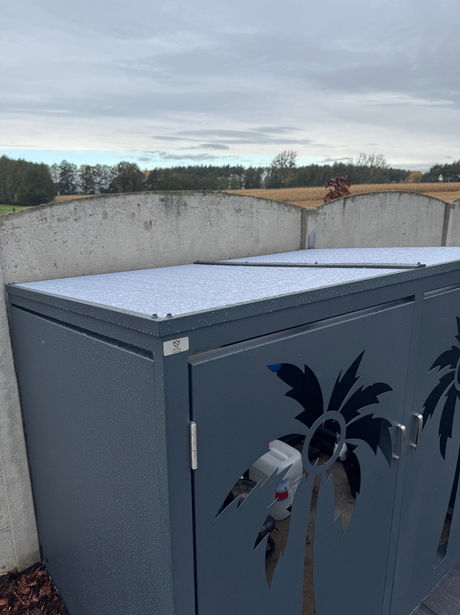 Metalowa obudowa na śmietniki z motywem palmy, szara, zamykana, stojąca przy betonowym ogrodzeniu. Widoczne krople deszczu.