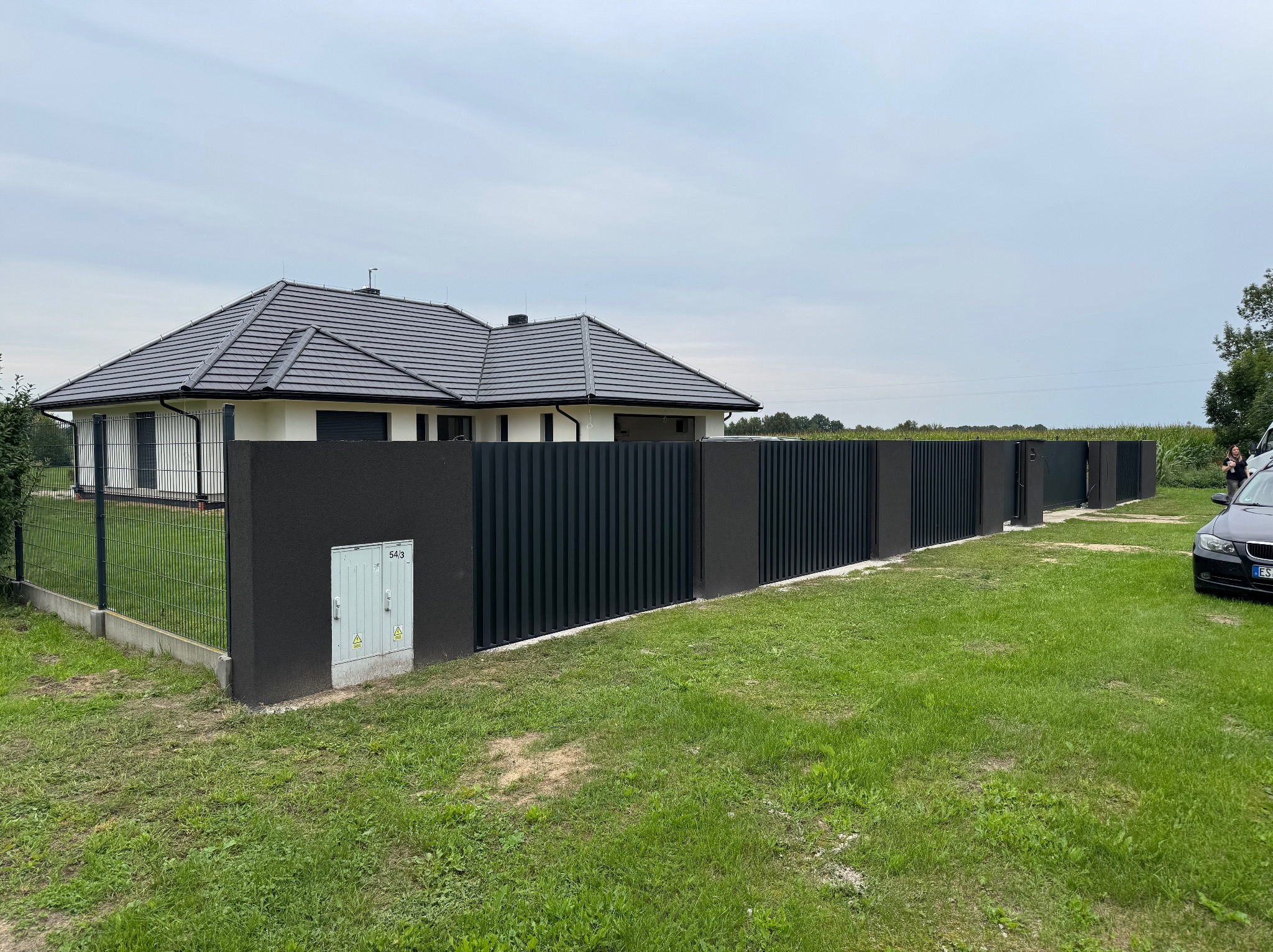 Nowoczesne ogrodzenie panelowe w kolorze antracytowym, z elementami murowanymi, otaczające dom jednorodzinny z ciemną dachówką. Widok z zewnątrz, w tle zielony trawnik i błękitne niebo.