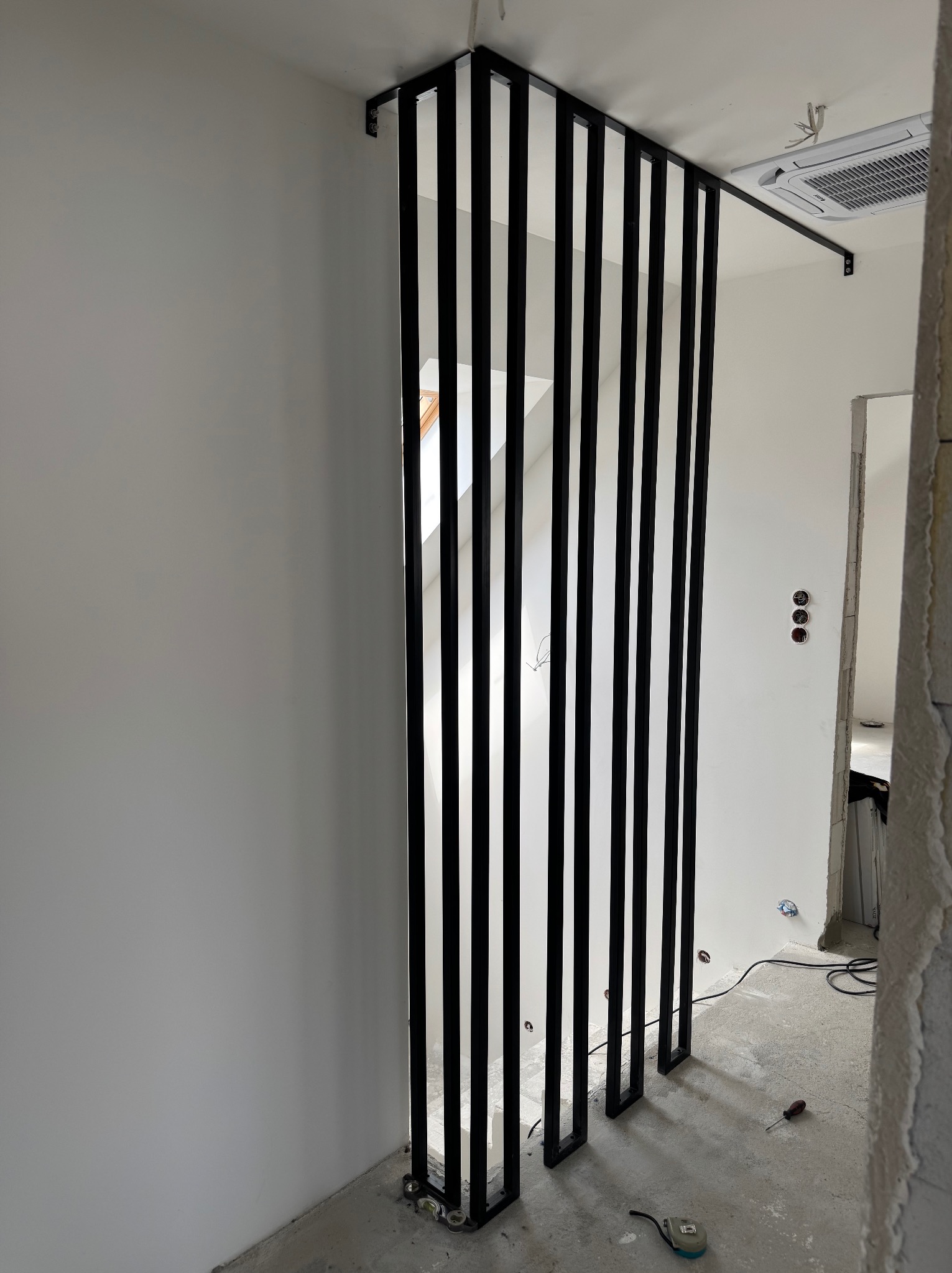 Nowoczesna, czarna ścianka działowa z metalowych profili, zamontowana w jasnym wnętrzu. Widoczne mocowania do sufitu i podłogi. Minimalistyczny design.