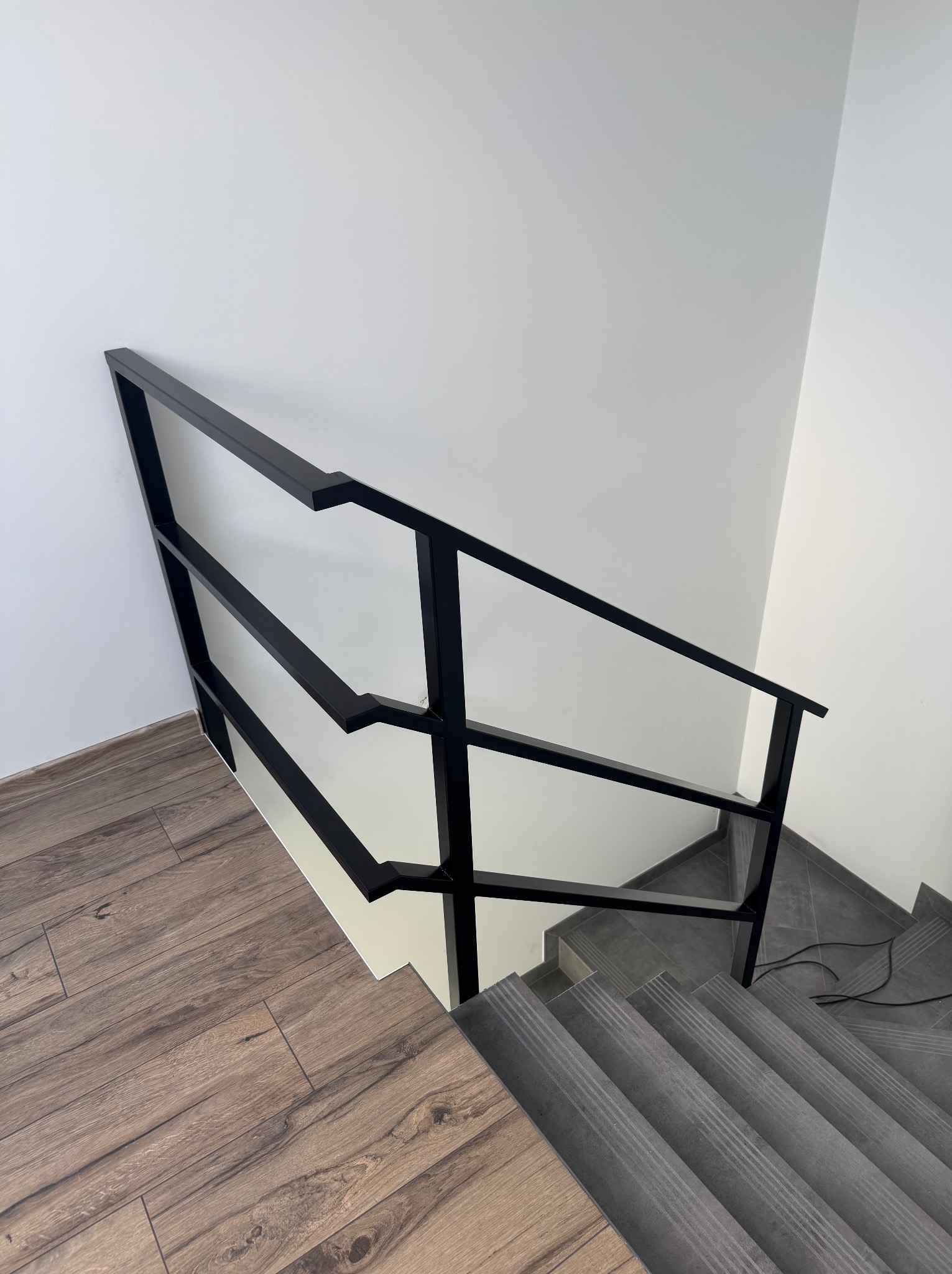 Minimalistyczna, czarna balustrada schodowa z metalu, widziana z góry. Kontrastujące połączenie drewnopodobnych paneli podłogowych i szarych płytek na schodach.