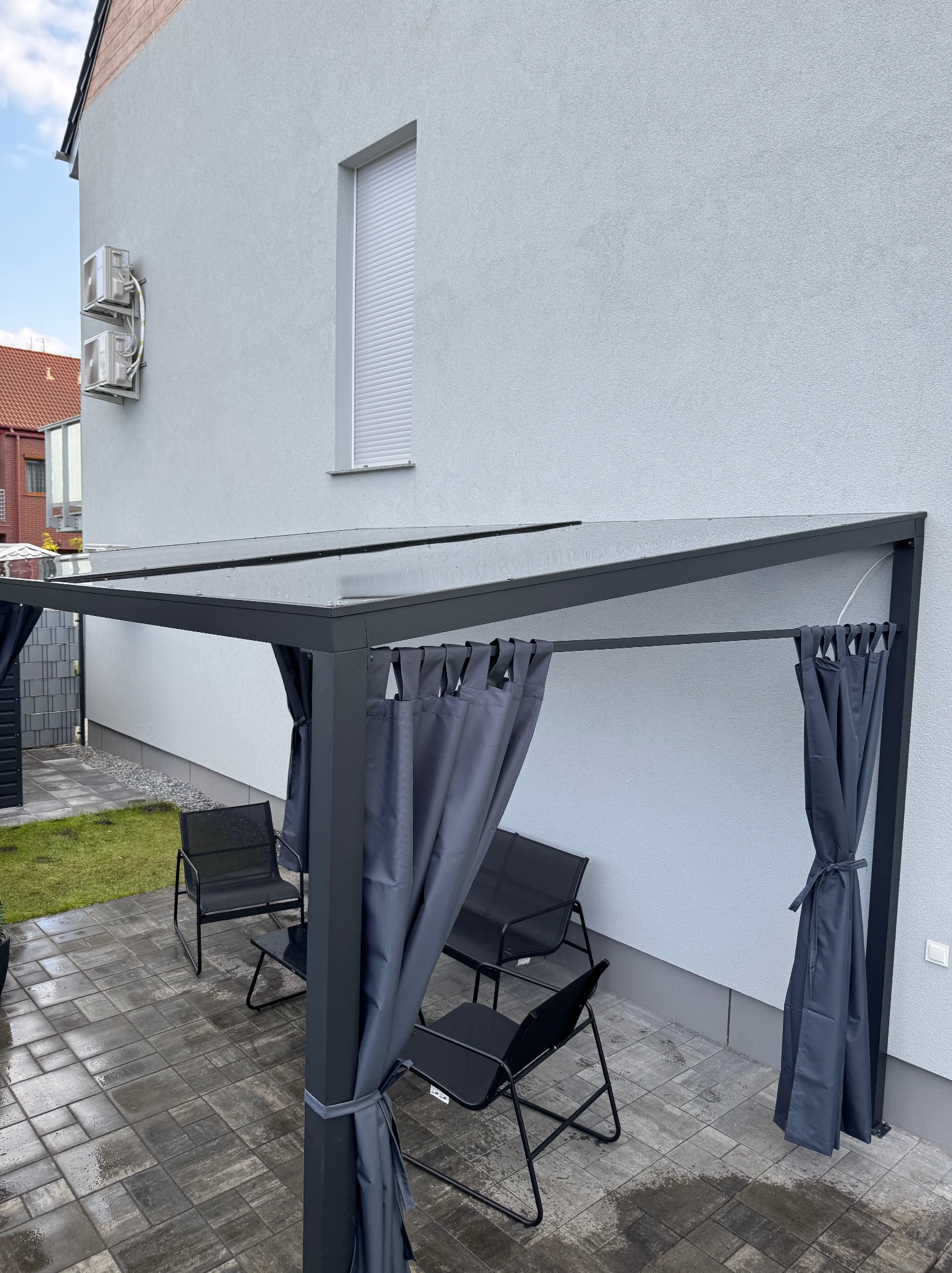 Nowoczesna pergola metalowa z zasłonami w Miliczu. Cztery czarne krzesła i stolik na szarej kostce brukowej. Jasnoszara ściana budynku w tle.