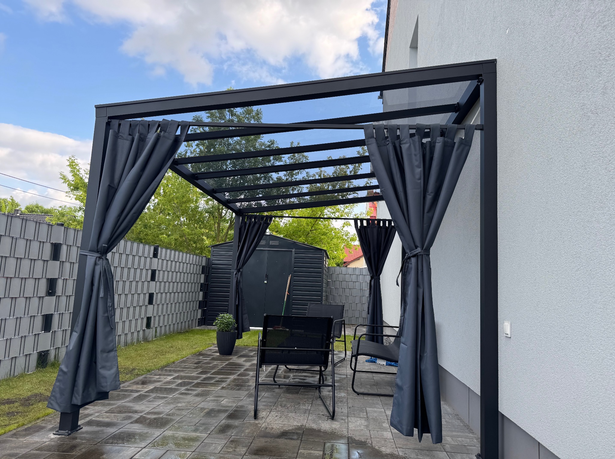 Nowoczesna, czarna pergola metalowa z zasłonami na tarasie. Widoczne meble ogrodowe i fragment budynku. Całość w stonowanej kolorystyce, zieleń w tle.