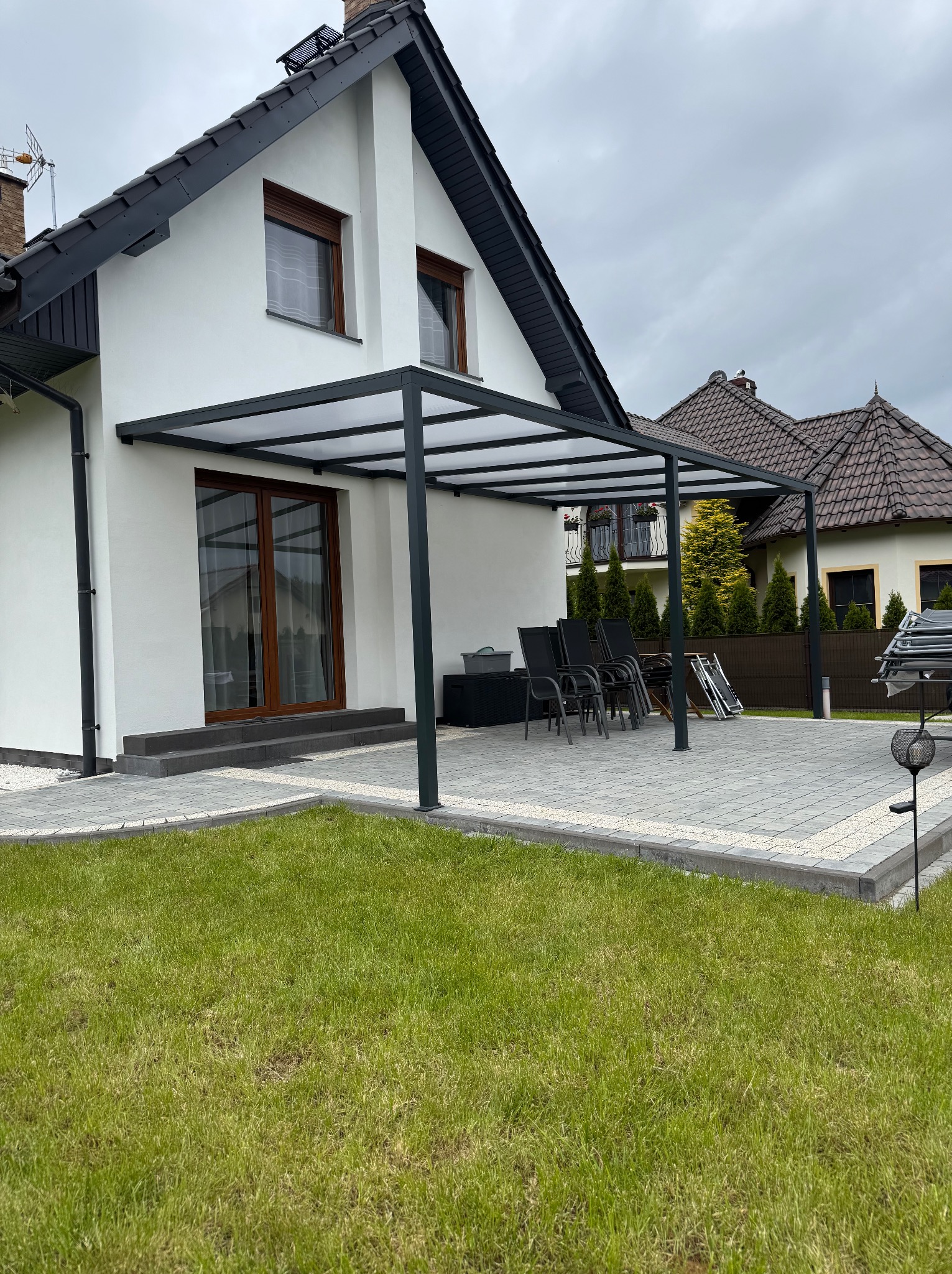 Nowoczesna pergola z ciemną ramą i przezroczystym zadaszeniem, przylegająca do białego domu z brązowymi oknami. Widoczne meble ogrodowe i trawnik.