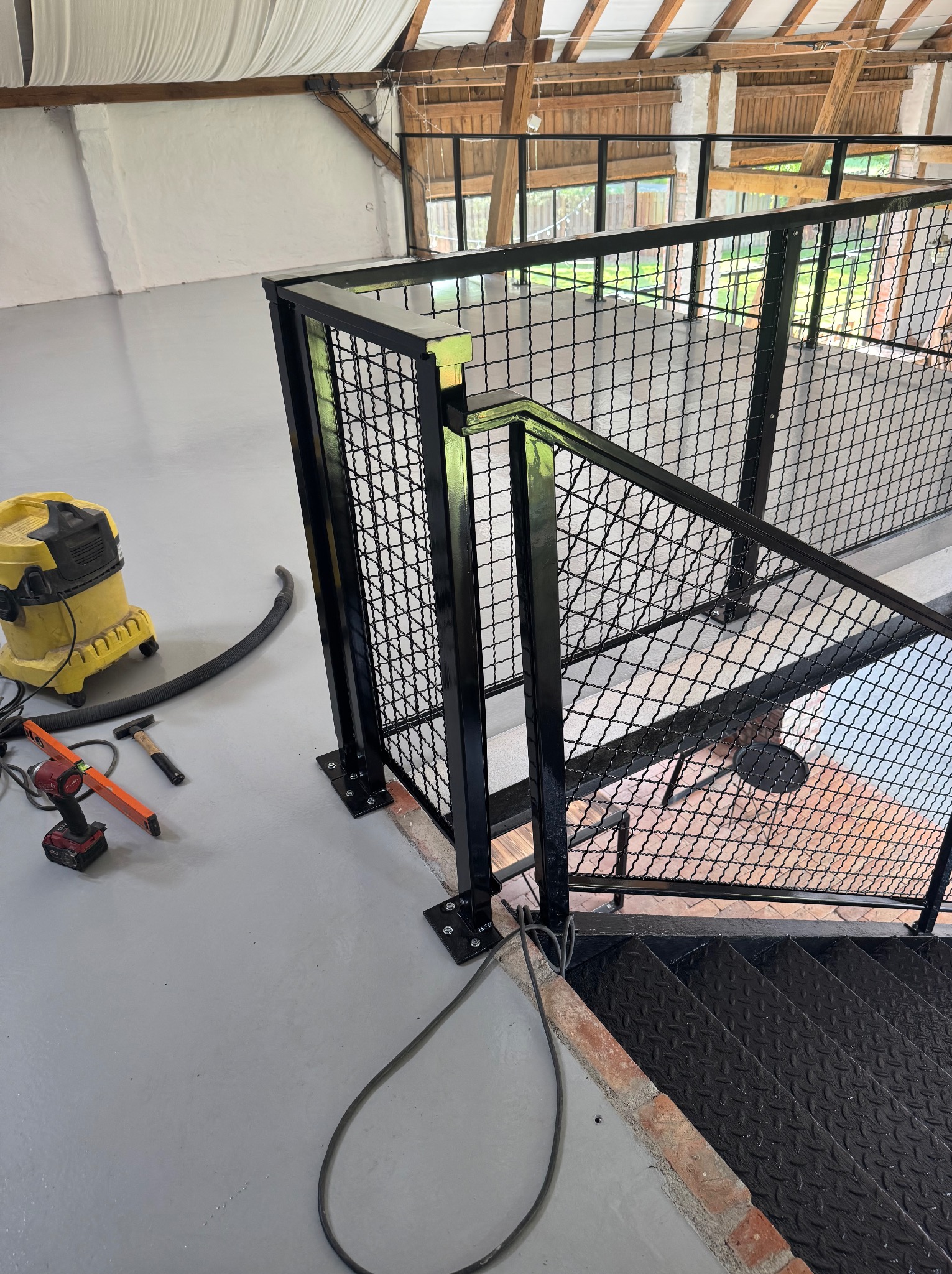 Czarna, metalowa balustrada z siatką na schodach w loftowym wnętrzu. Widoczne narzędzia i odkurzacz, sugerujące etap montażu. Surowy styl, cegła i drewno w tle.