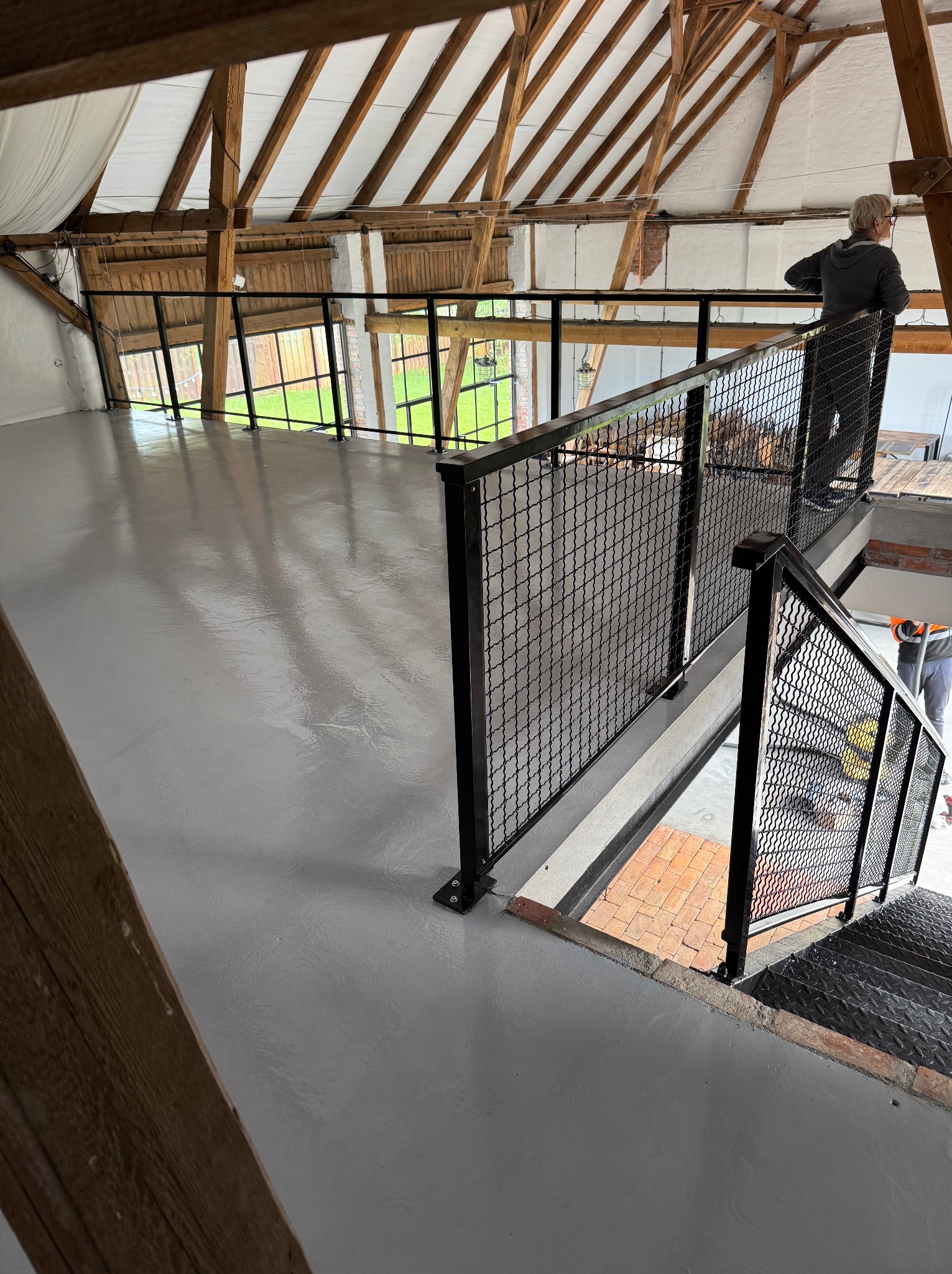 Antresola z metalową balustradą z siatki w loftowym wnętrzu. Widoczna osoba opierająca się o balustradę i schody. Drewniana konstrukcja dachu.