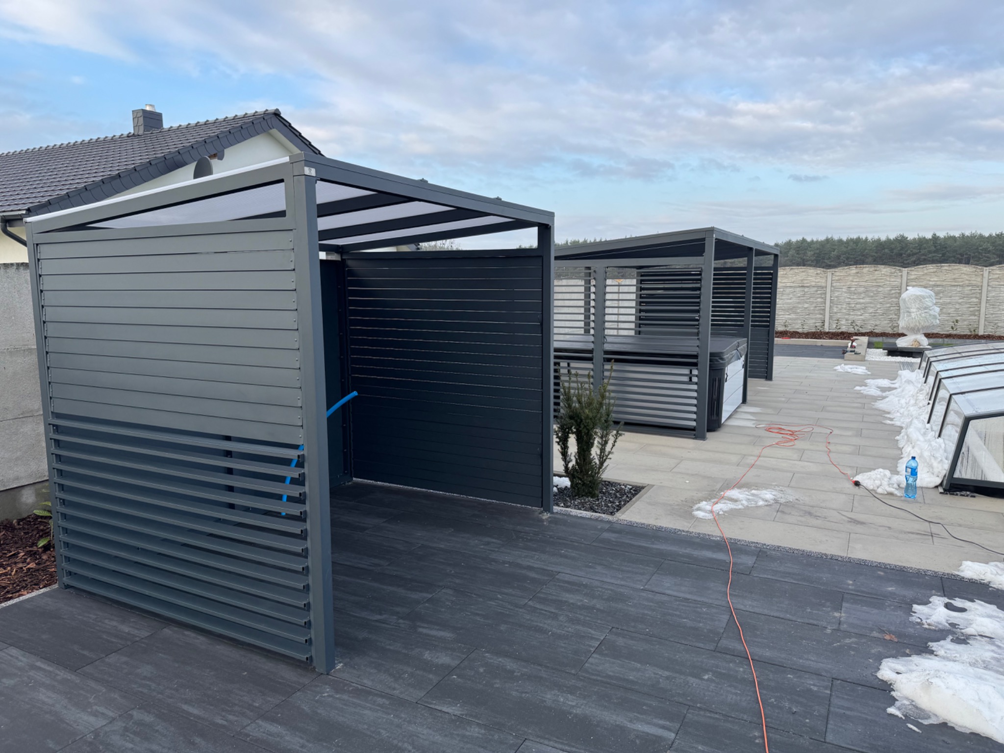 Dwie nowoczesne, szare pergole z lamelami i przezroczystym zadaszeniem, ustawione na szarym bruku. W tle dom z ciemną dachówką i biały płot. Częściowo stopniały śnieg.