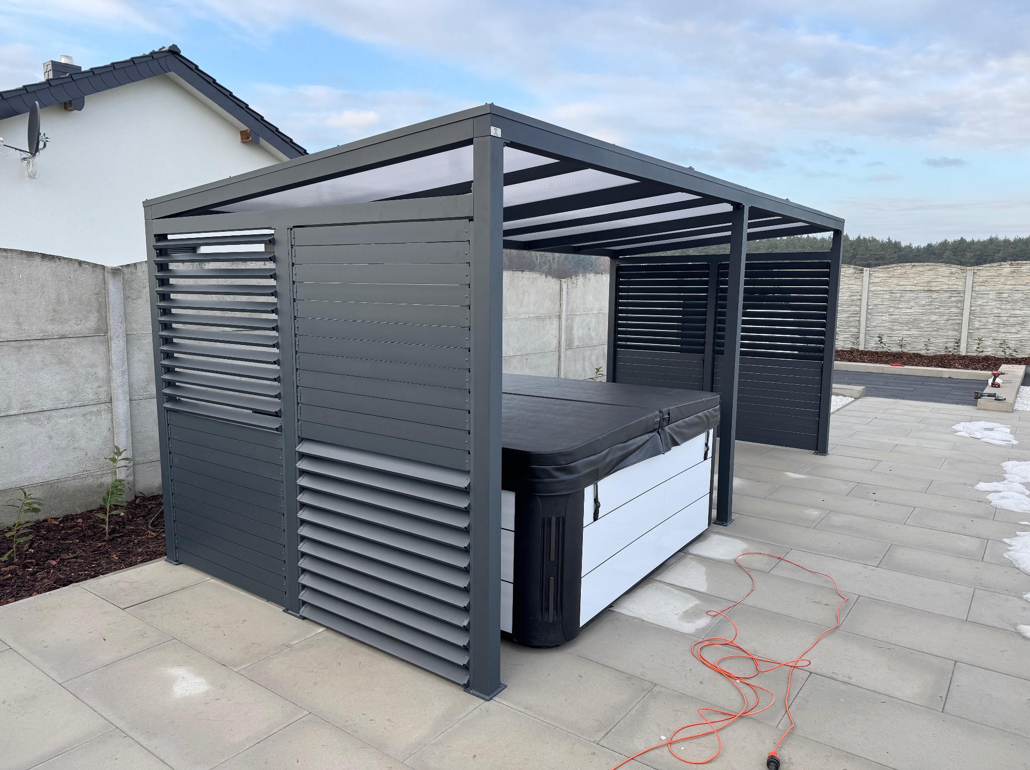 Nowoczesna, szara pergola z lamelami osłaniająca jacuzzi. Widoczne elementy konstrukcyjne i detale wykończenia. Tło stanowi betonowy płot i fragment domu.