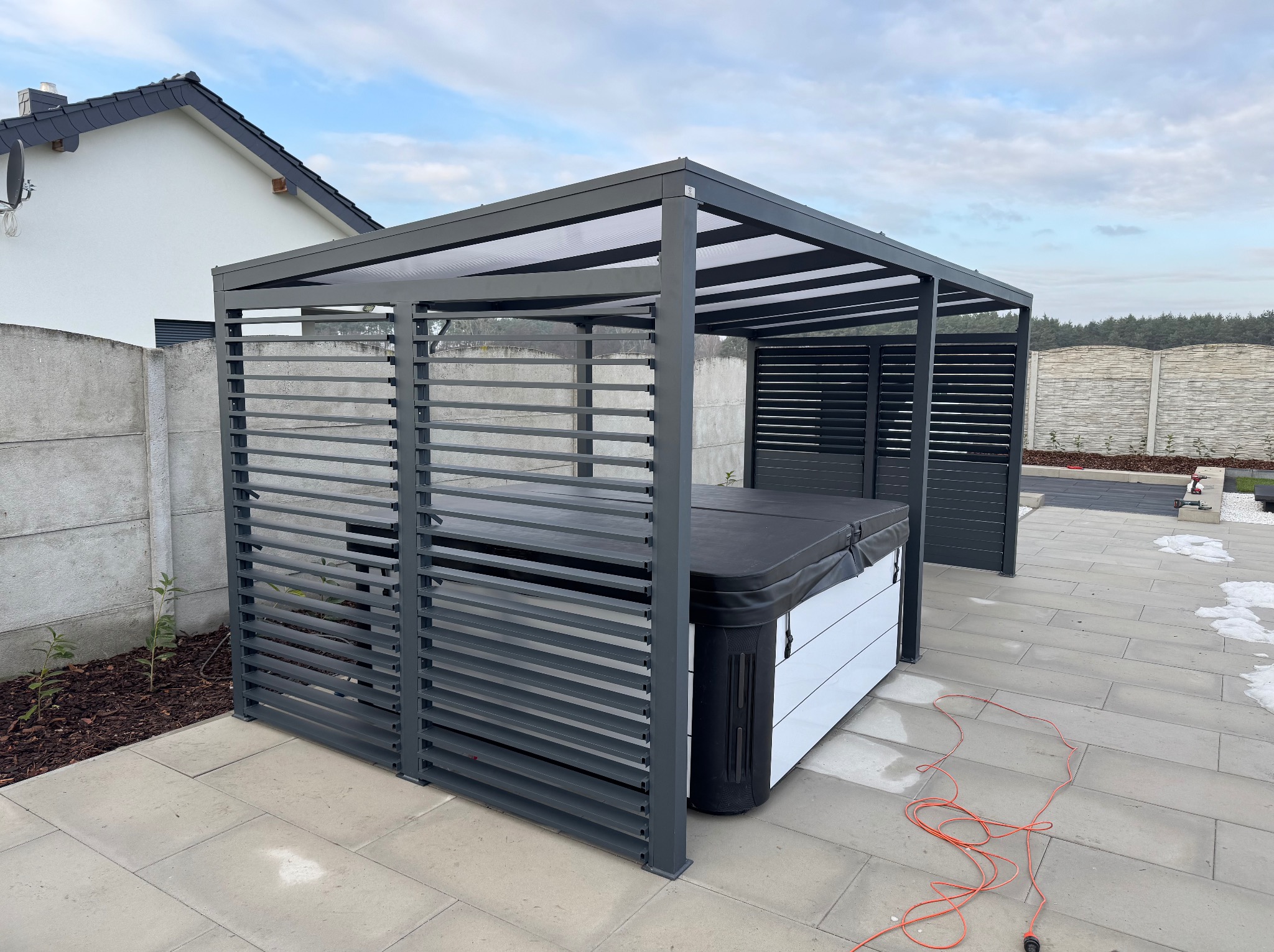 Nowoczesna, szara konstrukcja z poziomych listew osłaniająca jacuzzi na zewnątrz. Widoczne elementy domu i ogrodzenia. Pomarańczowy kabel na kostce brukowej.