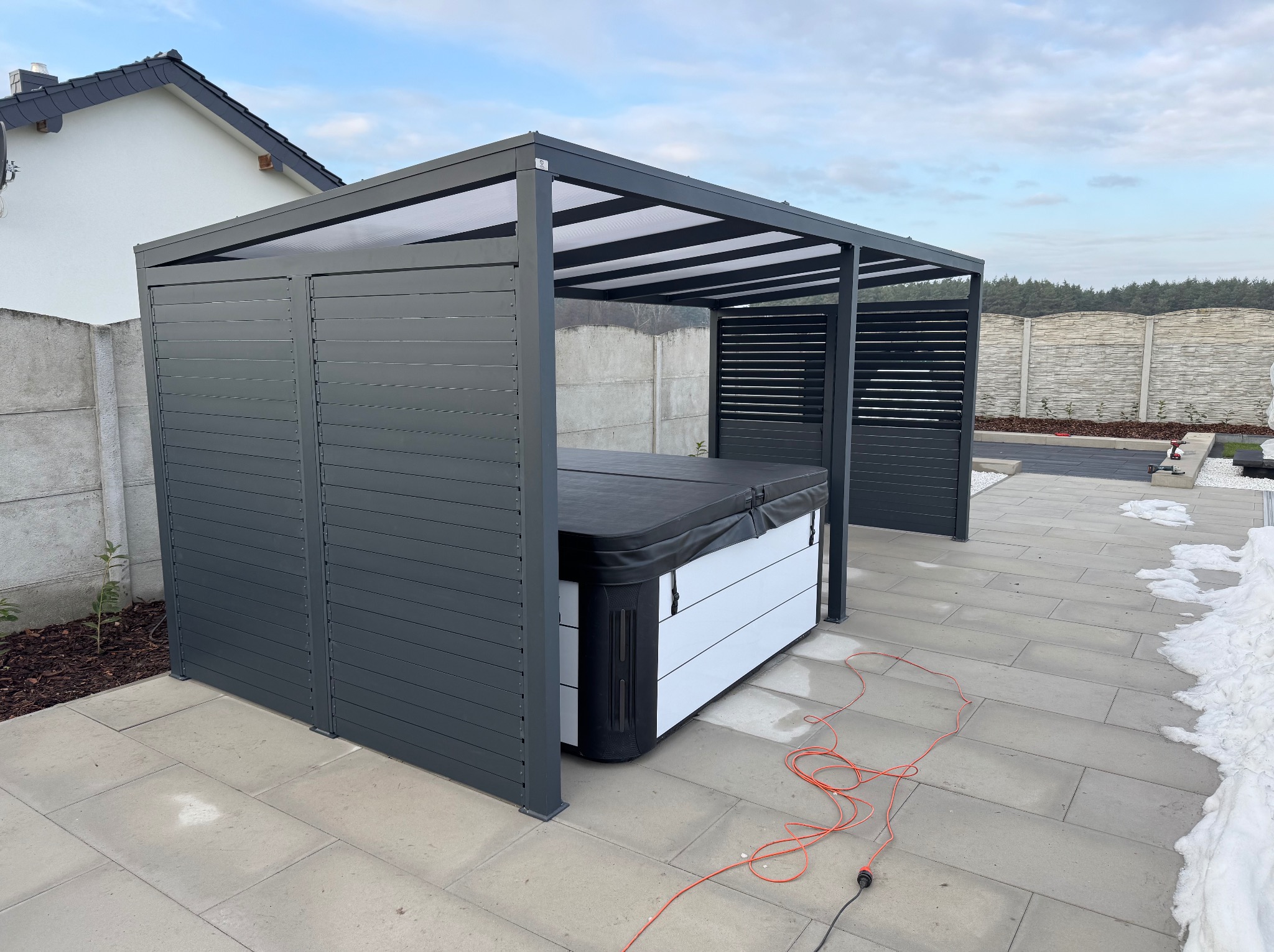 Nowoczesna, szara pergola metalowa z częściową zabudową, osłaniająca jacuzzi. Widoczne panele boczne i zadaszenie z prześwitującego materiału. Na pierwszym planie kabel zasilający.