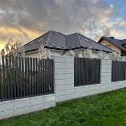 DAJUX SPÓŁKA Z OGRANICZONĄ ODPOWIEDZIALNOŚCIĄ - Nowoczesne ogrodzenie z paneli pionowych w kolorze grafitowym, osadzone na betonowych słupkach z dekoracyjnymi trawami, na tle domu z ciemnym dachem i zachmurzonego nieba o zachodzie słońca.