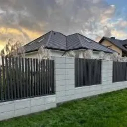 Nowoczesne ogrodzenie z paneli pionowych w kolorze grafitowym, osadzone na betonowych słupkach z dekoracyjnymi trawami, na tle domu z ciemnym dachem i zachmurzonego nieba o zachodzie słońca.