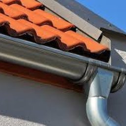 DAJUX SPÓŁKA Z OGRANICZONĄ ODPOWIEDZIALNOŚCIĄ - Nowy, błyszczący system rynnowy z metalu, zamontowany na ścianie budynku z pomarańczową dachówką ceramiczną, widoczny na tle błękitnego nieba.