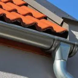 Nowy, błyszczący system rynnowy z metalu, zamontowany na ścianie budynku z pomarańczową dachówką ceramiczną, widoczny na tle błękitnego nieba.