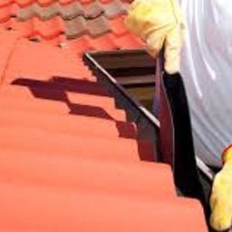 DAJUX SPÓŁKA Z OGRANICZONĄ ODPOWIEDZIALNOŚCIĄ - Dekarz w żółtych rękawicach montuje rynnę na dachu pokrytym czerwoną dachówką.