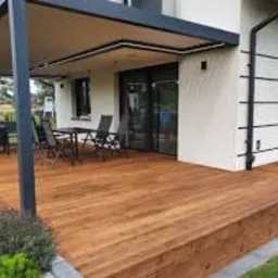 Drewniany taras z pergolą przy nowoczesnym domu jednorodzinnym, widok na meble ogrodowe i zadbany trawnik.