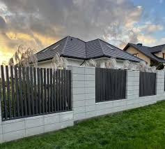 Nowoczesne ogrodzenie z paneli pionowych w kolorze grafitowym, osadzone na betonowych słupkach z dekoracyjnymi trawami, na tle domu z ciemnym dachem i zachmurzonego nieba o zachodzie słońca.