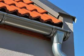 Nowy, błyszczący system rynnowy z metalu, zamontowany na ścianie budynku z pomarańczową dachówką ceramiczną, widoczny na tle błękitnego nieba.