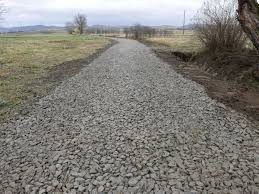 Droga gruntowa wysypana szarym tłuczniem, przygotowana pod ułożenie kostki brukowej na tle łąki i drzew bez liści.