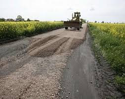 Równiarka wyrównuje drogę gruntową na tle kwitnącego pola rzepaku, widoczne świeże ślady po pracy maszyny.