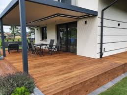 Drewniany taras z pergolą przy nowoczesnym domu jednorodzinnym, widok na meble ogrodowe i zadbany trawnik.