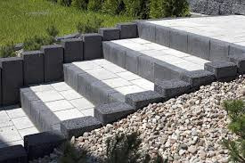 Schody zewnętrzne wyłożone jasnymi płytami i obramowane ciemnoszarą kostką, z dekoracyjnym podsypaniem z drobnych kamieni i zielenią w tle.