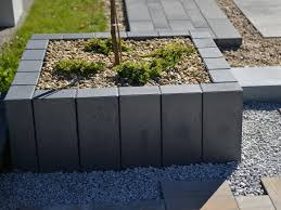 Kwadratowa donica z szarej kostki brukowej otaczająca małe drzewko i krzewy, wysypana drobnymi kamieniami, fragment ścieżki z kostki brukowej w tle.